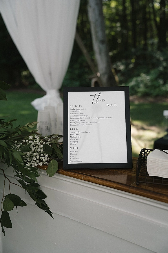 Bar menu sign in a black frame on a rustic wood bar top with greenery garland and baby's breath at an outdoor garden setup