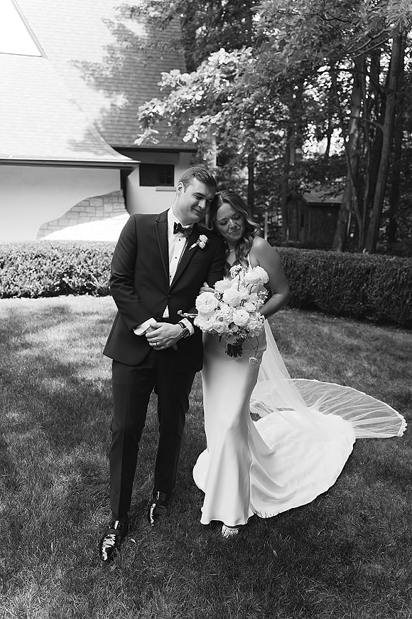 Couple portrait in a black and white wedding portrait, bride and groom hugging with bouquet and long veil on a garden lawn near a house exterior