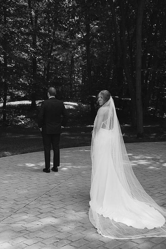 Wedding first look as bride in a wedding dress and long veil approaches tuxedoed groom on a woodland patio in dappled sunlight