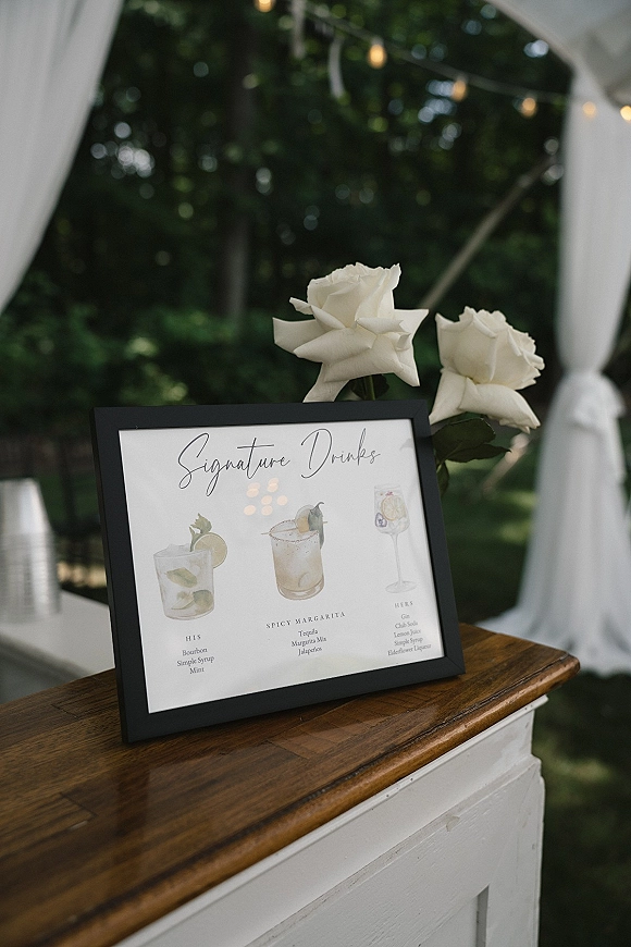 Signature drinks sign in a black frame with cocktail illustrations and white roses on a wood bar, set beneath string lights and drapery outdoors