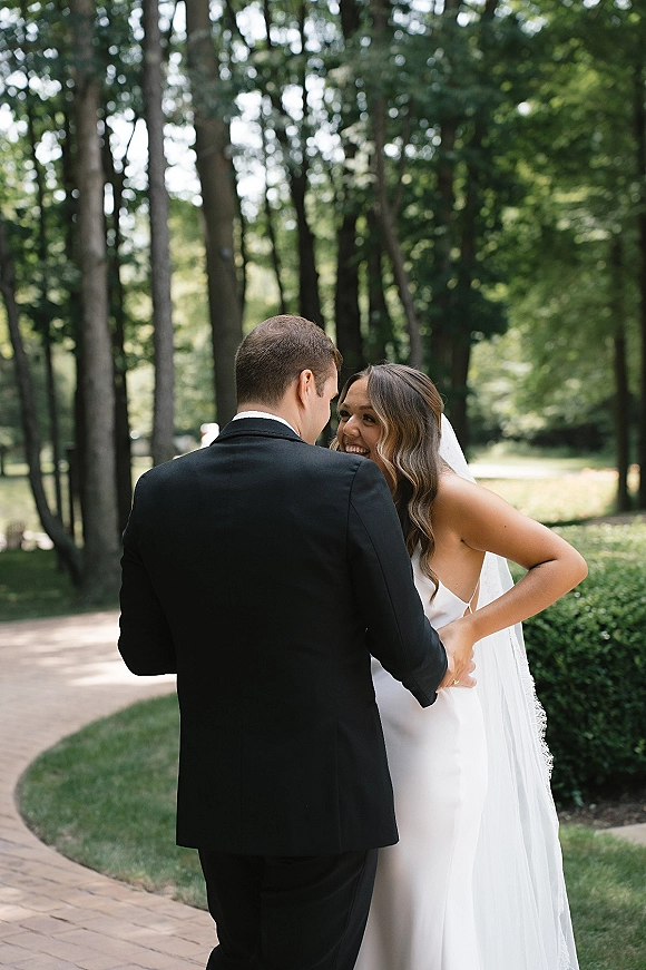 Wedding couple portrait of bride and groom laughing while holding hands on a garden path, her long veil flowing behind his tuxedo