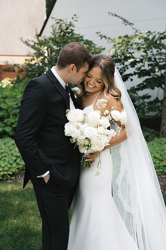 Couple portrait of bride and groom embracing as he kisses her forehead, bride holding a white rose bouquet in a lush garden setting