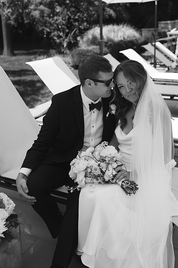 Couple portrait of bride and groom seated as he kisses her cheek, bouquet and veil visible, on poolside lounge chairs under shade