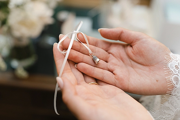 Wedding keepsake charm and bridal bouquet charm held in manicured hands with a white ribbon and silver pendant, lace sleeve and ring, blurred indoor florals behind