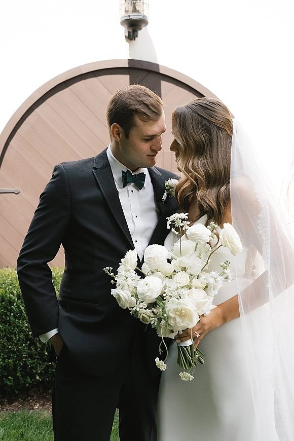 Couple portrait of bride and groom holding bouquet, her veil draped over strapless dress, beside an arched doorway with lantern and greenery hedge
