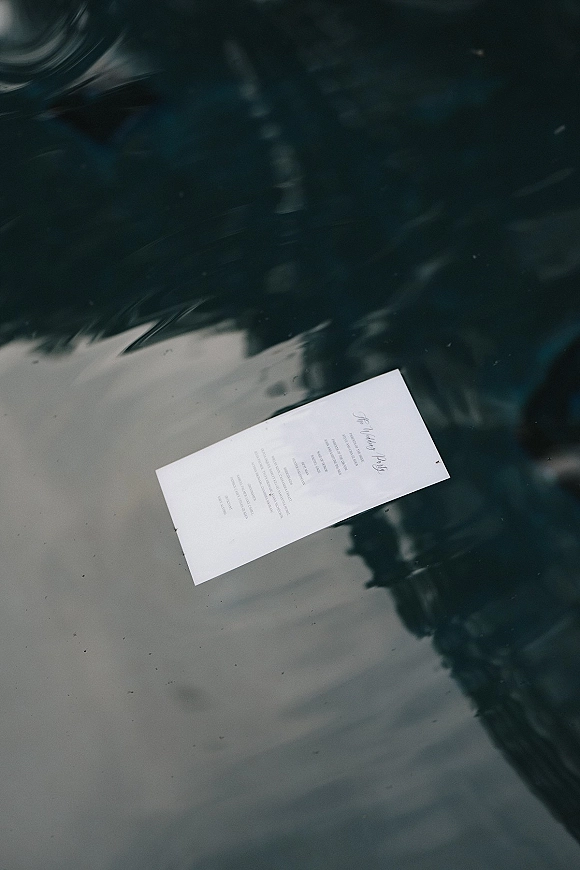 Wedding invitation flat lay with calligraphy printed text on a reflective glass surface, with subtle greenery reflections in the background