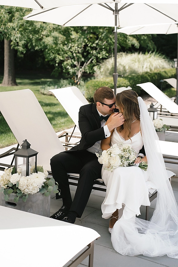 Couple portrait of groom kissing bride’s forehead as they sit on lounge chairs, bride holding white bouquet on a garden patio lawn
