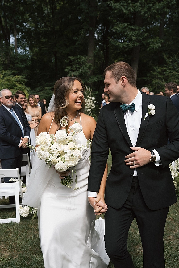 Wedding recessional as bride and groom walk down aisle holding hands, bride with white bouquet and veil, guests in garden setting