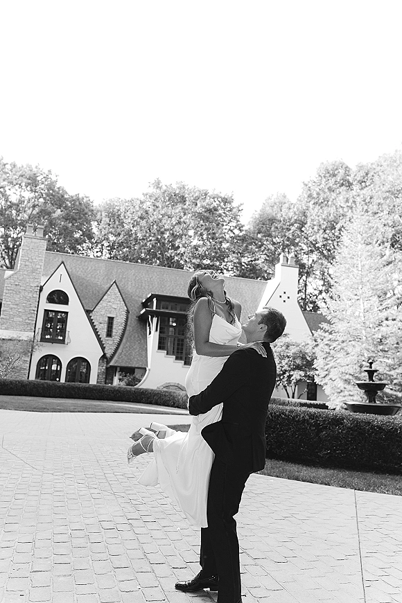 Couple portrait of groom lifting bride in a playful wedding photo, her strapless dress and heels by a fountain at an estate driveway