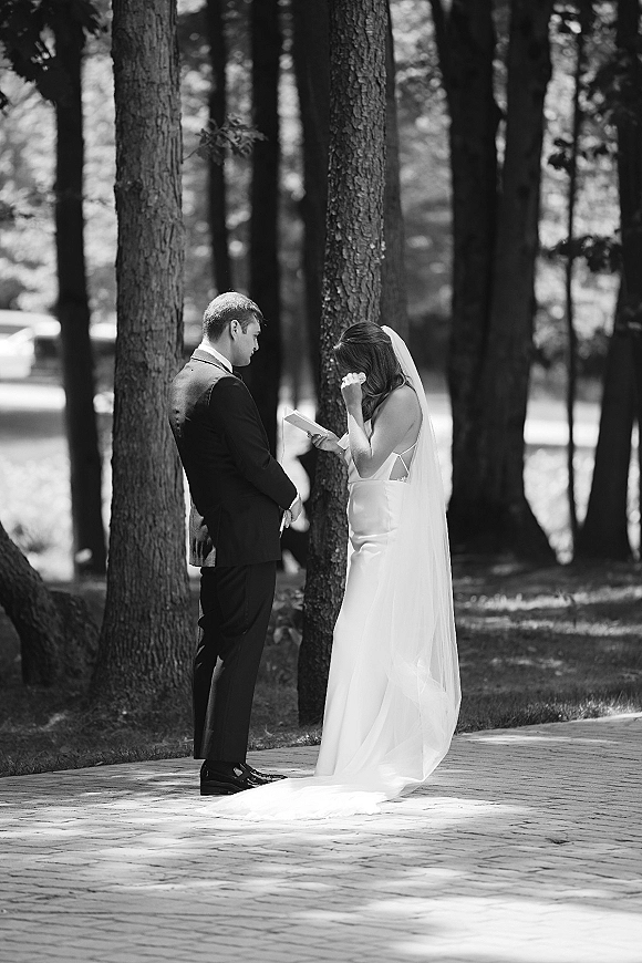 Wedding vows as bride in veil reads from a vow book while groom in tuxedo listens on a tree-lined walkway in dappled sunlight