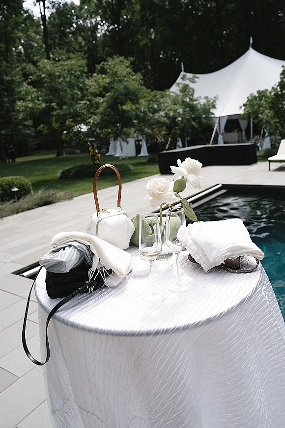 Wedding details on a poolside table with white roses in glass vases, wine glasses, mini handbags, and a towel beneath string lights by a tent