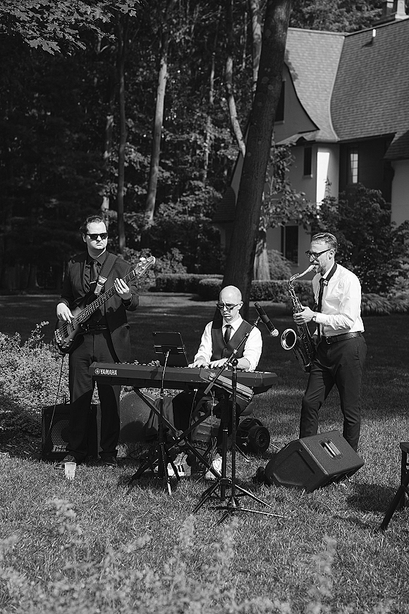 Wedding band playing live wedding music with saxophone, keyboard and bass on a grassy lawn near trees and a house exterior