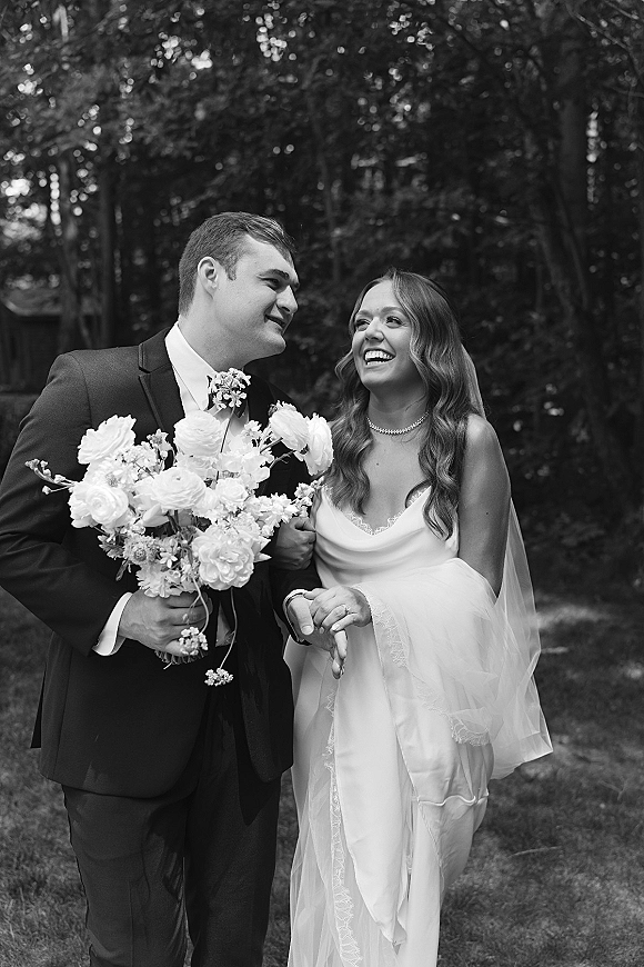 Couple portrait, black and white wedding photo of bride and groom laughing, holding hands and bouquet on a tree-lined garden lawn