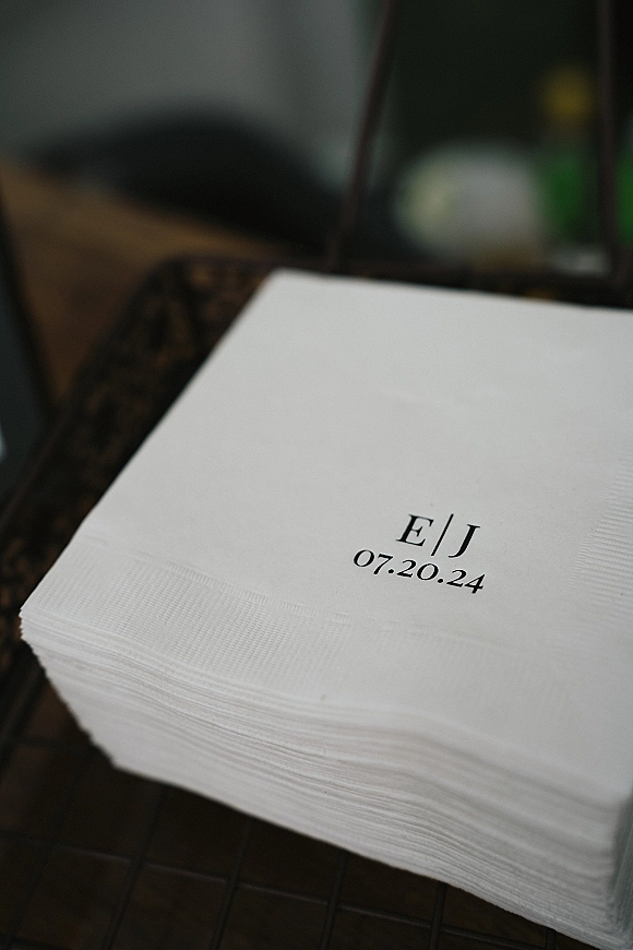 Wedding cocktail napkins with custom wedding napkins monogram and date printing beside a beverage glass on a dark tabletop