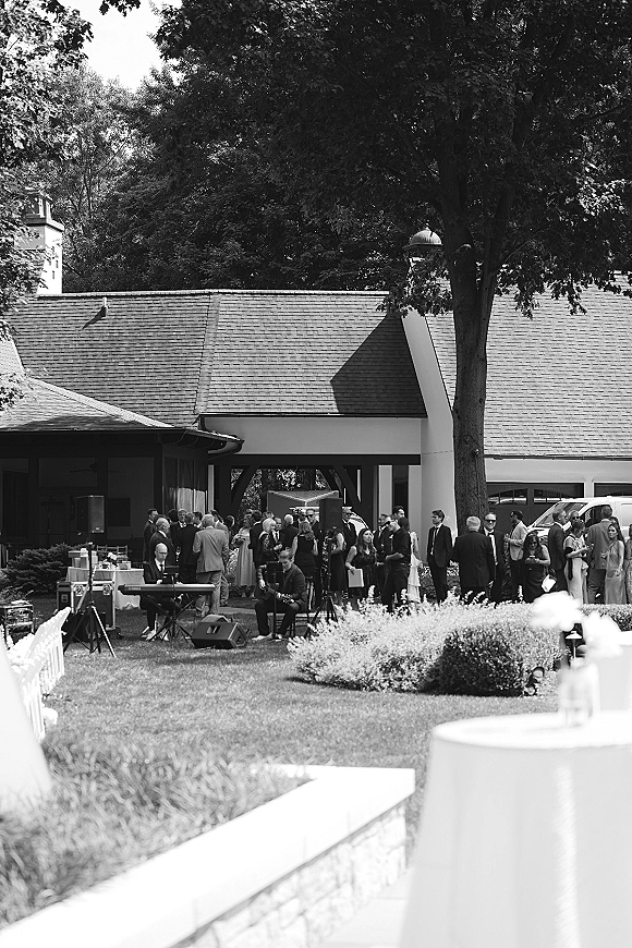 Wedding cocktail hour with guests mingling on a lawn while a live band plays near white linen cocktail tables under a large tree