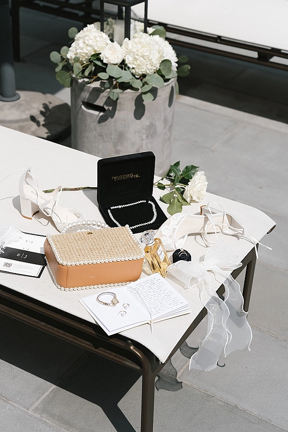 Wedding details flat lay of bridal accessories with invitations, white heels, rings, pearl clutch and roses on a gray tiled outdoor patio