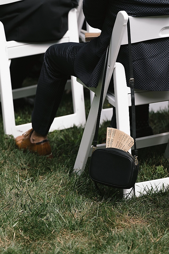 Wedding guest detail with a black crossbody purse and lace hand fan beside white folding chairs on a grass lawn outdoor seating area