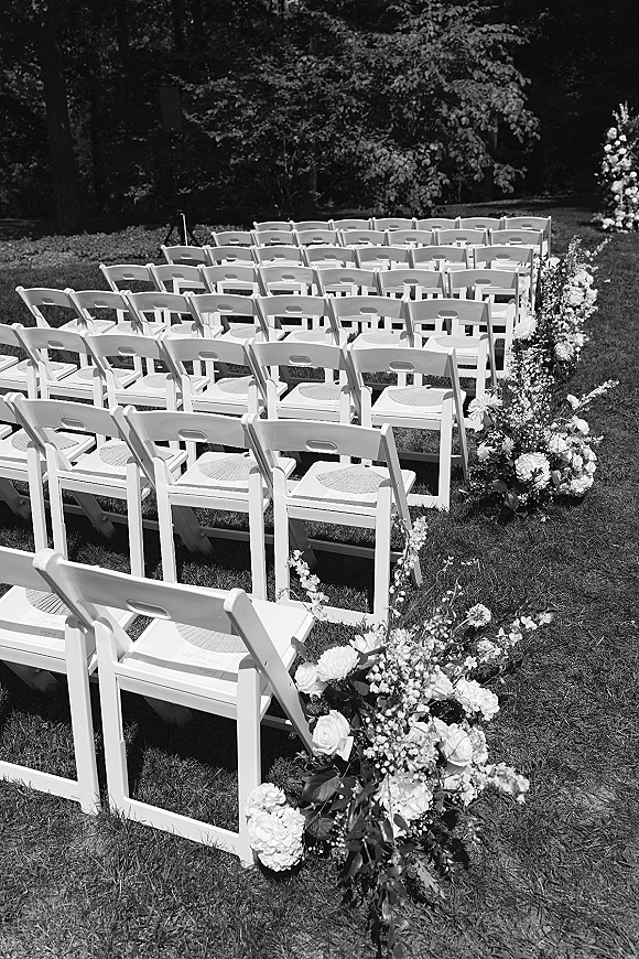 Ceremony seating with white folding chairs lining a floral-edged aisle on a grass lawn, framed by trees in a garden setting