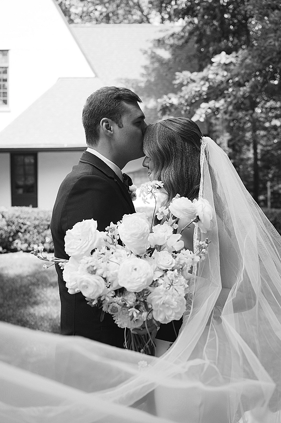 Wedding couple portrait in black and white as bride and groom embrace with a forehead kiss, her veil draped over a white bouquet outdoors