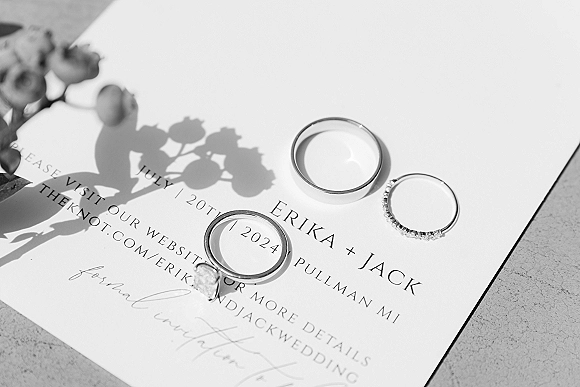 Wedding rings flatlay with engagement ring close up atop a wedding invitation and floral sprig on a light stone surface