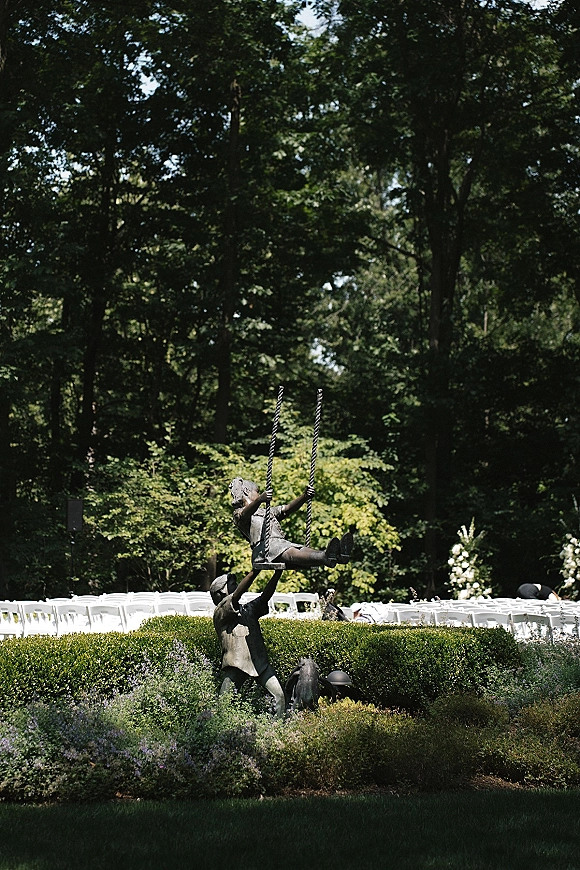 Outdoor ceremony seating with rows of white folding chairs on a garden lawn, with a statue and rope swing among hedges and trees