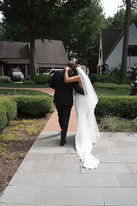 Newlywed couple walking down a garden path, bride holding groom’s shoulder as her long veil and dress train flow behind them