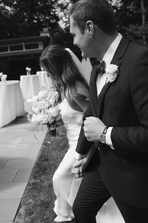 Couple portrait of bride and groom walking hand in hand, bride holding a white bouquet with veil trailing on an outdoor patio with greenery