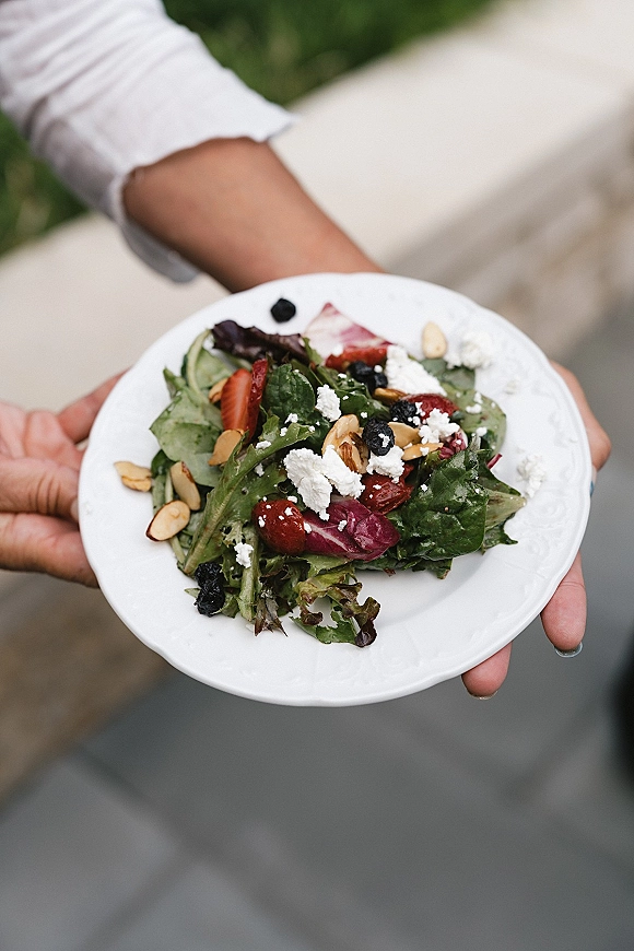 Wedding salad course with mixed greens, strawberries, blueberries, sliced almonds, and crumbled cheese on a white plate held outdoors near greenery
