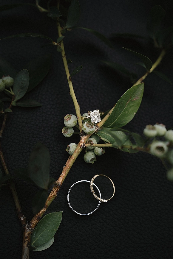 Wedding rings in a wedding ring flatlay with an emerald-cut diamond engagement ring, stacked bands, and greenery on dark fabric backdrop