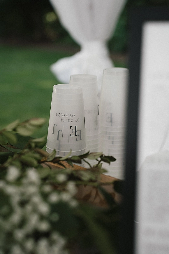 Wedding favor cups stacked on a wooden tray with greenery garland and white flowers, set before a white drape on a lawn