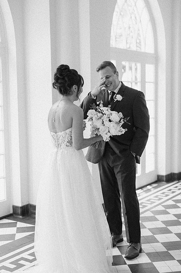 Wedding first look as groom turns to see bride in a strapless lace dress holding a bouquet in an arched-window hallway with tile floor