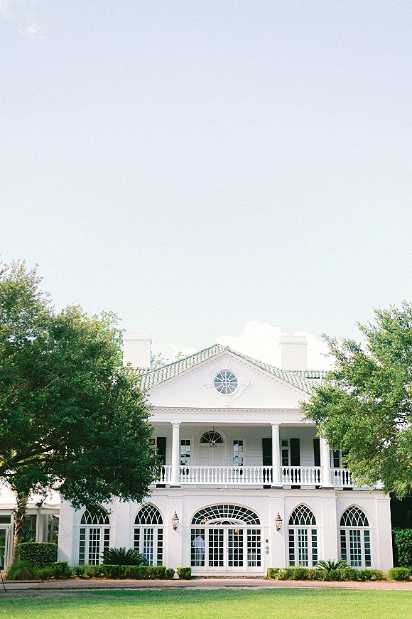 Wedding venue exterior with arched windows, white mansion wedding venue facade featuring French doors, balcony and columns framed by trees and lawn