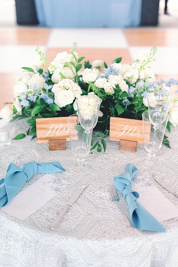 Reception tablescape with white and blue floral centerpiece, wood place cards, blue napkins, and champagne flutes on patterned cloth outdoors