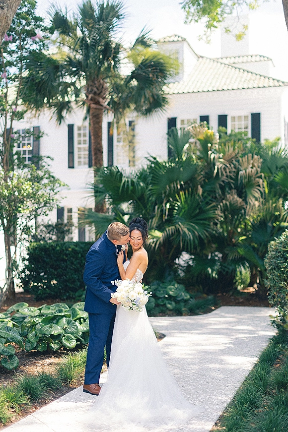Couple portrait of groom kissing bride’s cheek as she holds a white and green bouquet, with veil and palm-lined garden walkway behind