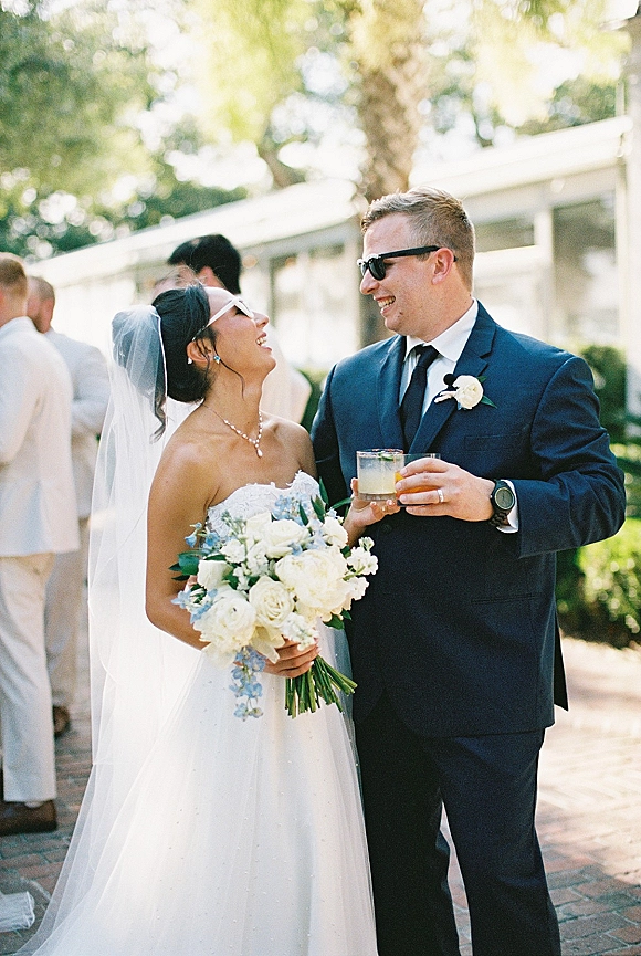 Couple portrait of bride and groom laughing in sunglasses, holding cocktail glasses as wedding guests mingle on a sunlit garden walkway