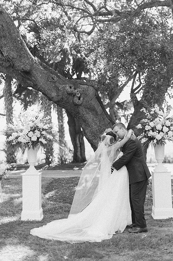 Wedding kiss portrait of bride and groom kissing, her long veil flowing over a strapless gown beside floral urns under a large tree