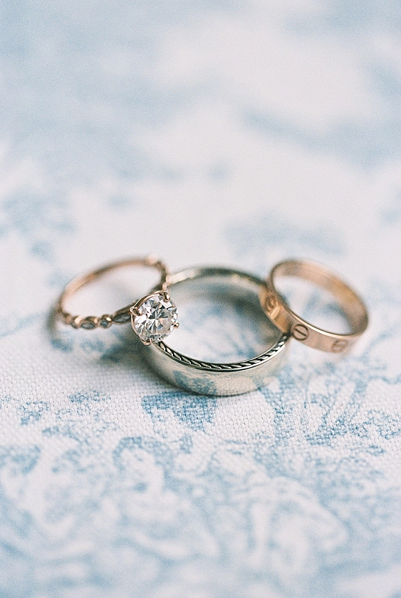 Wedding rings close up featuring a diamond engagement ring beside gold and silver bands with braided detail on light blue patterned fabric