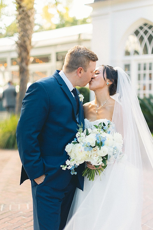 Wedding kiss portrait of bride and groom kissing, her veil flowing as she holds a blue and white bouquet outside a white building with arches