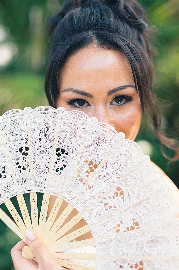 Bridal portrait of a bride holding lace fan over her face, showcasing soft makeup and updo hairstyle against lush garden foliage backdrop