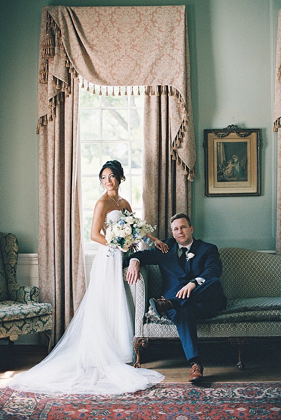 Couple portrait of bride holding bouquet beside seated groom in navy suit, lit by window light in an elegant room with drapes