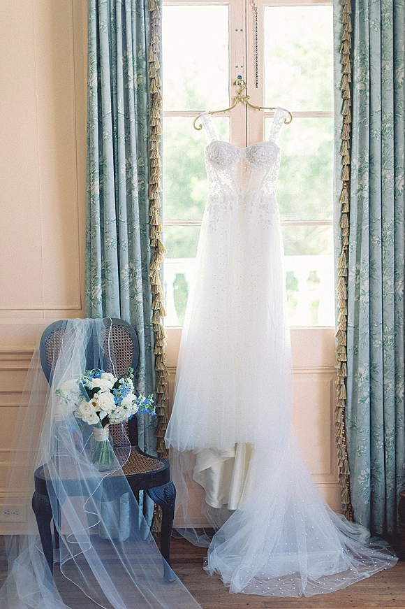 Wedding dress hanging in a window, strapless lace bodice with sweetheart neckline, veil and blue-and-white bouquet near floral curtains