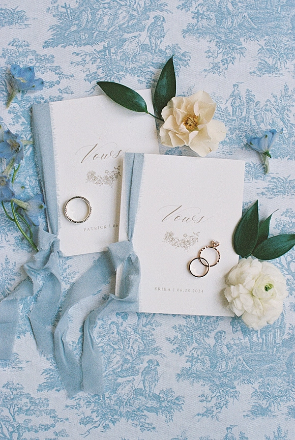 Wedding vow books tied with ribbon beside wedding rings, styled with flowers and greenery on blue toile patterned fabric flat lay