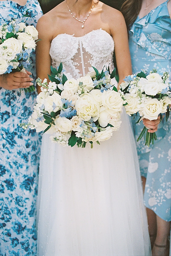 Bridesmaid bouquets of white roses and pale blue blooms tied with greenery, held by the wedding party in blue floral dresses and pearls