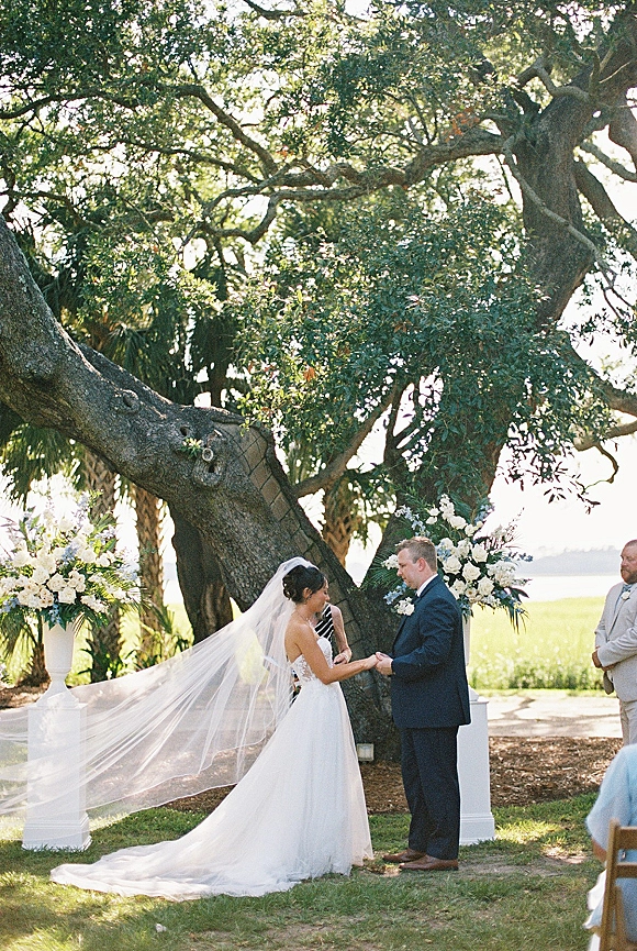 Wedding vows during an outdoor wedding ceremony as bride and groom hold hands beneath an oak tree by the waterfront, veil drifting behind her