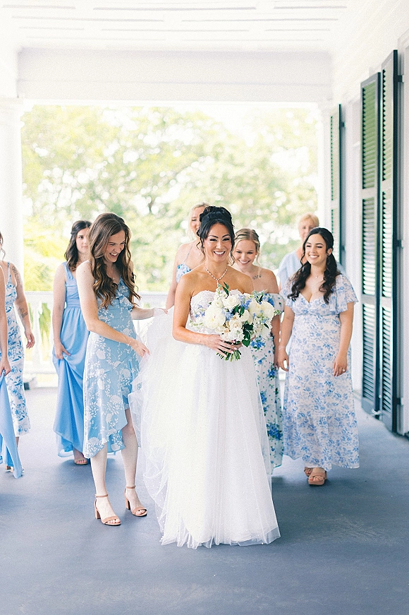 Bride with bridesmaids walking together, bride holding a white rose bouquet in a strapless tulle dress on a covered porch with green shutters