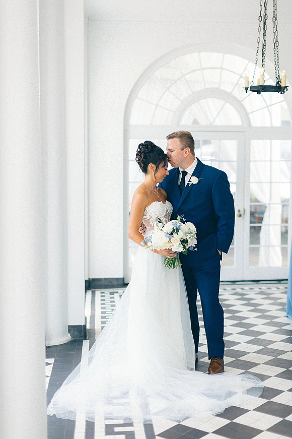 Couple portrait of groom kissing bride’s forehead as they embrace, her white bouquet and lace train flowing in a columned hall on checkered floor