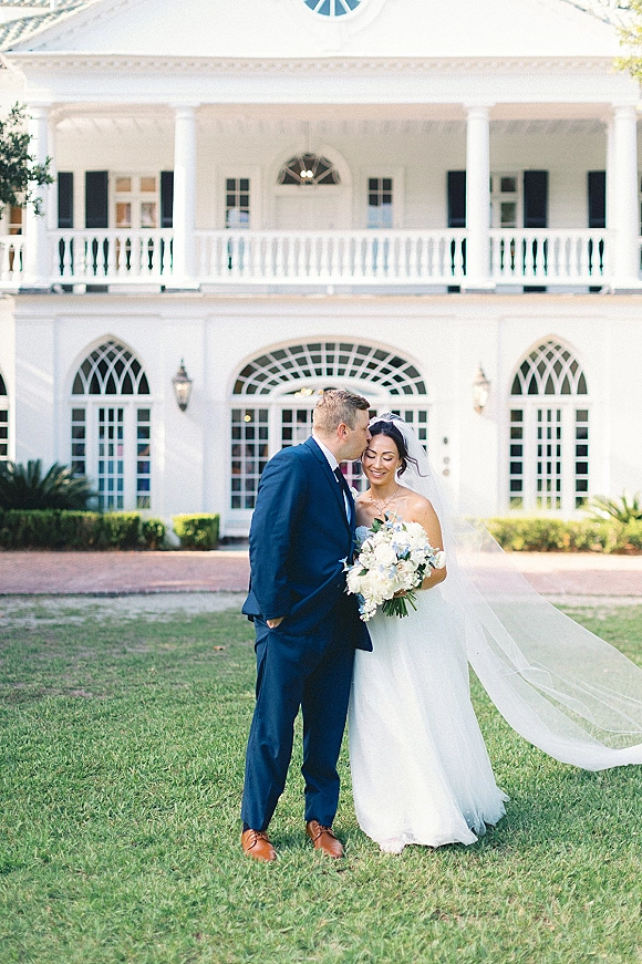 Couple portrait of groom kissing bride’s forehead as she holds a white and blue bouquet, veil blowing before a grand white mansion facade