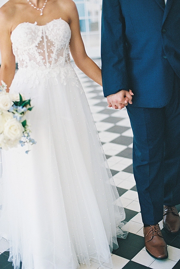 Couple holding hands as they walk, bride’s lace gown and bouquet visible over a black and white checkered floor in bright interior