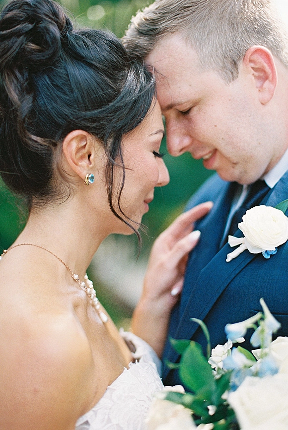 Couple portrait with a forehead touch as the bride in a strapless gown holds a bouquet beside groom with white rose boutonniere in greenery