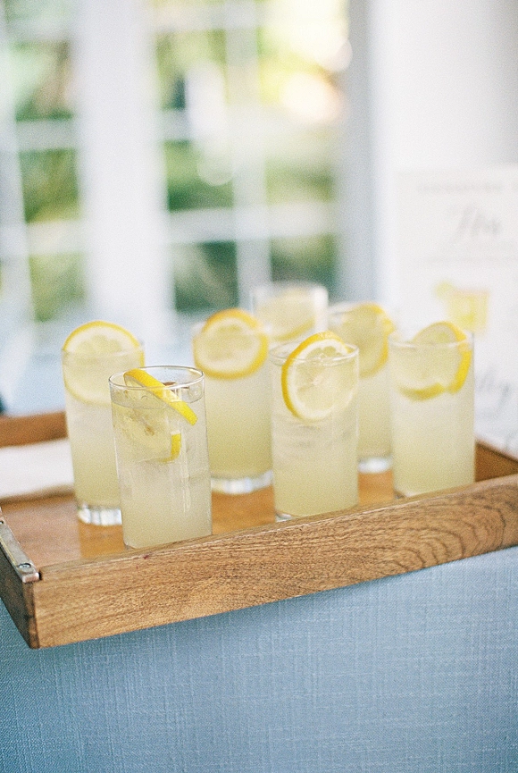 Wedding drinks in clear glasses with ice and lemon slice garnish on a wooden tray by window panes in soft indoor light